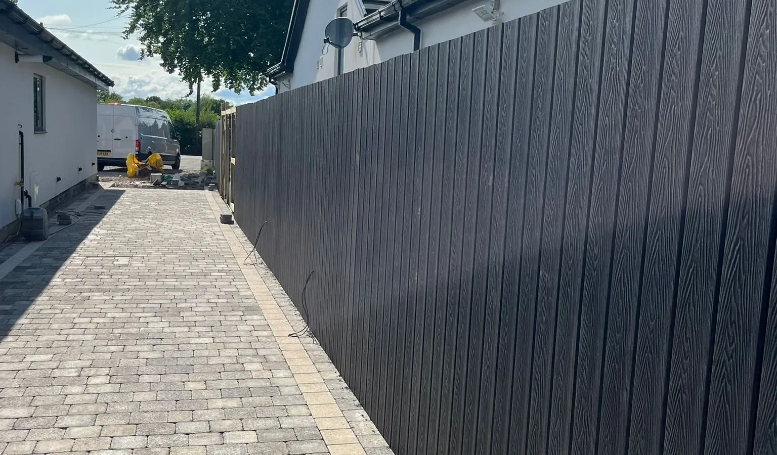 Block paved pathway with dark composite fence beside white rendered bungalow under blue sky