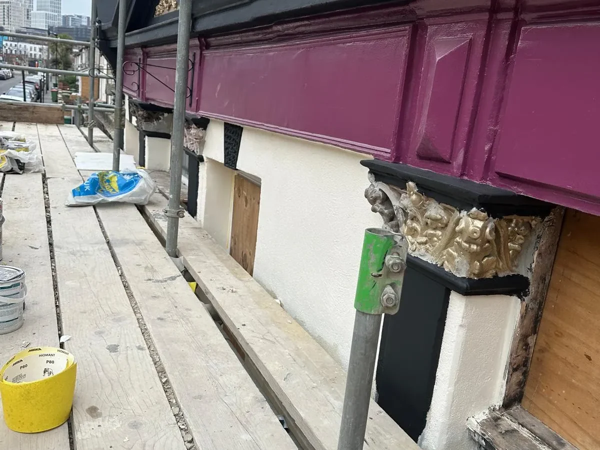 Victorian shopfront facade restoration with painted magenta cornice and gilded corbels under scaffolding